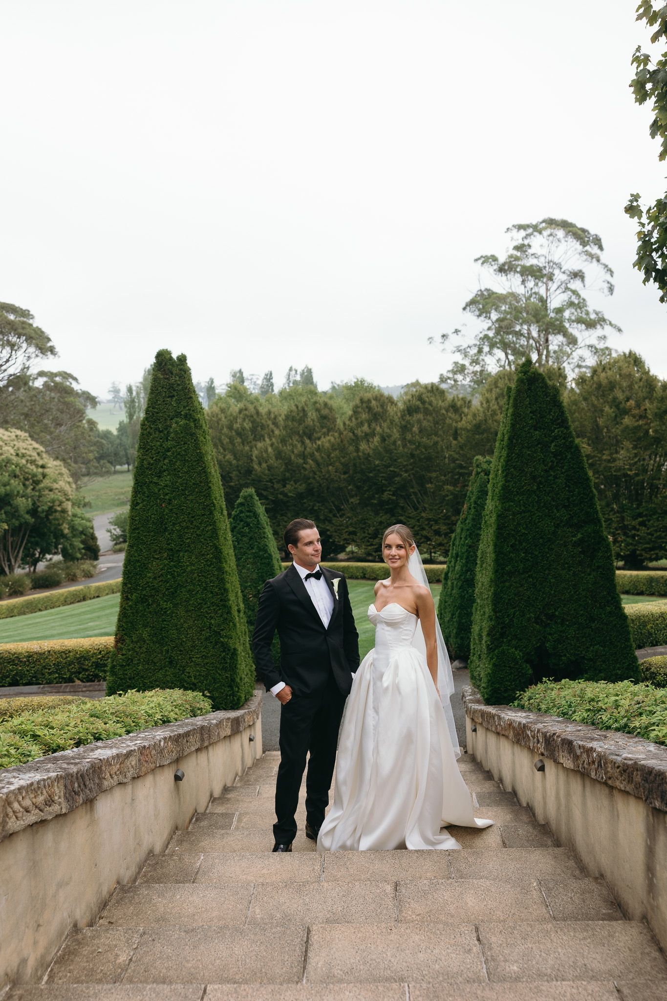 Bride and groom walk hand in hand through elegant formal garden with tall cypress trees on their wedding day.