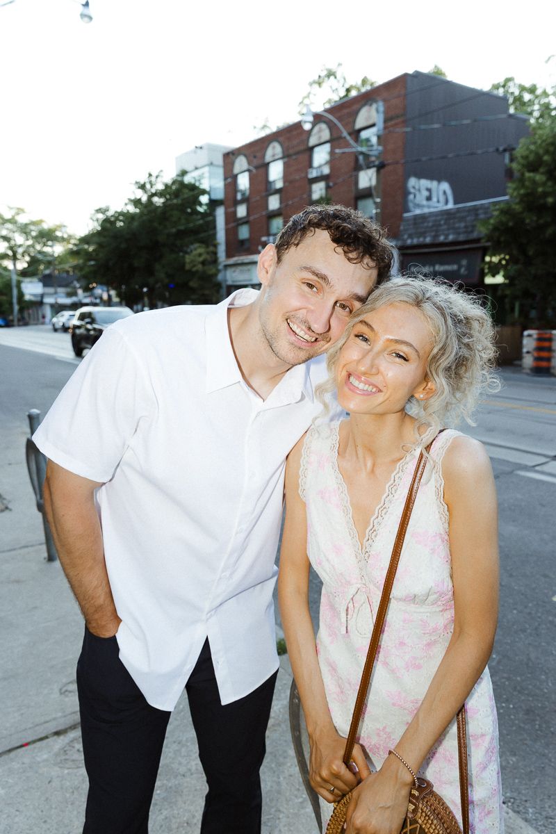 A smiling couple poses together on a city street, man in white shirt and woman in floral dress with crossbody bag.
