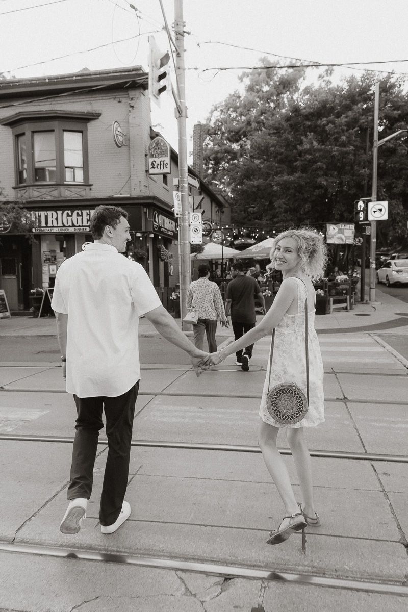 Black and white photo of a couple holding hands on a city street corner, woman in dress with round bag, man in white shirt.