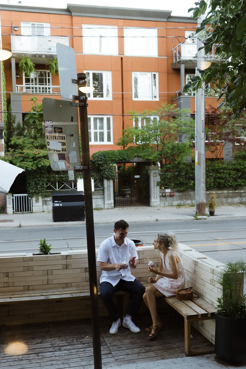 A couple sits on a wooden bench outside a café, chatting on a sunny urban street with an orange apartment building behind.
