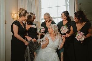 Bride in white gown sits surrounded by bridesmaids in black dresses holding pink bouquets during wedding preparations  at Boyne Hill House