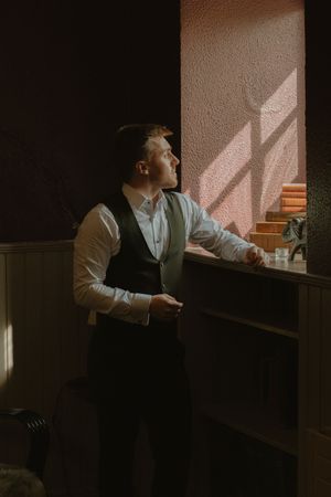 Groom stands by a sunlit window in a dimly lit room at Boyne Hill House Estate, Co. Meath,