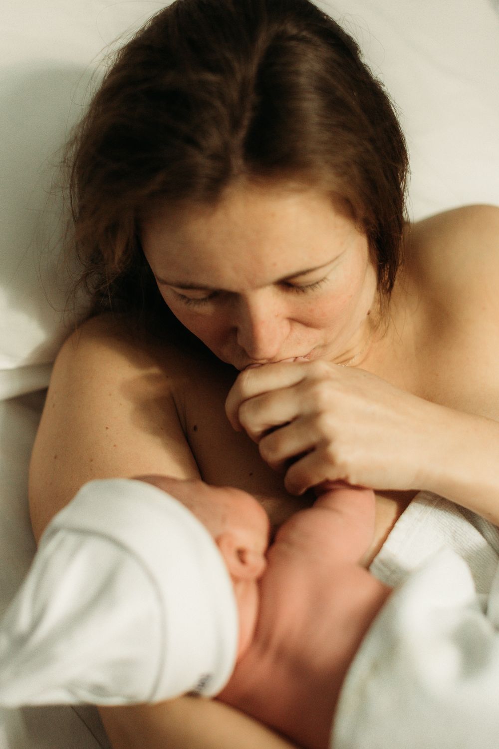 A new mother tenderly kisses her newborn baby's hand while lying down, wrapped in a white blanket.