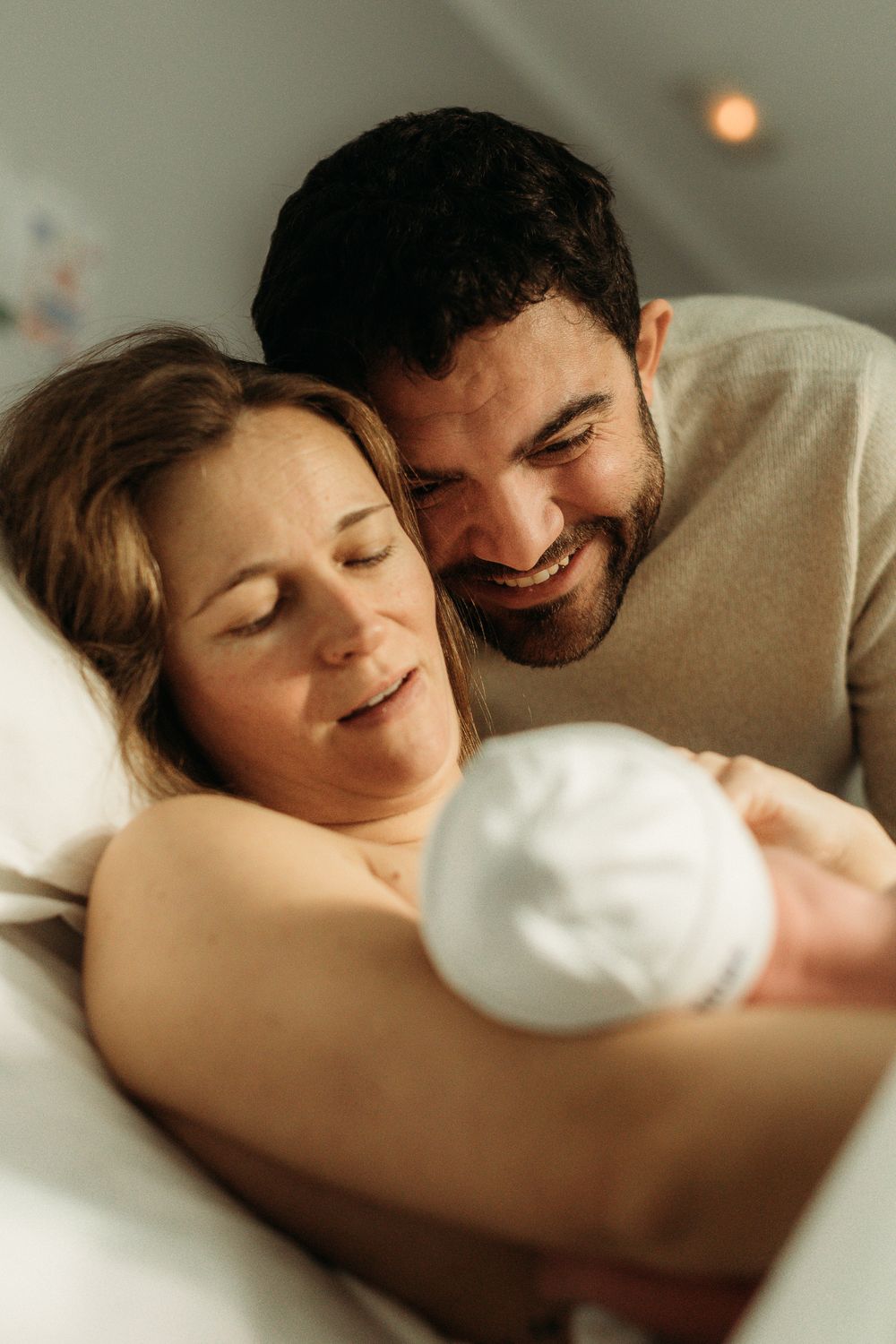 Happy couple smiling at their newborn baby wrapped in white, sharing a tender moment in a hospital bed.