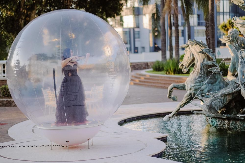 Person in elegant gown inside large transparent inflatable bubble sphere near decorative horse fountain outdoors.