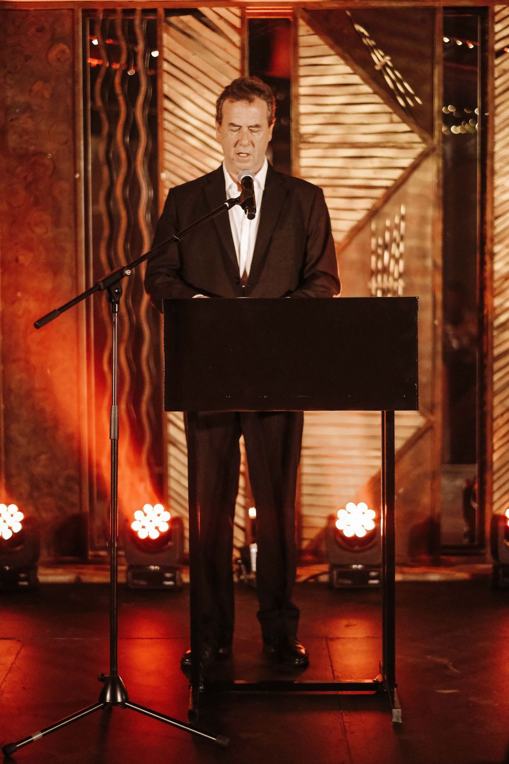 Man in tuxedo speaking at podium on stage with dramatic red lighting and decorative backdrop.