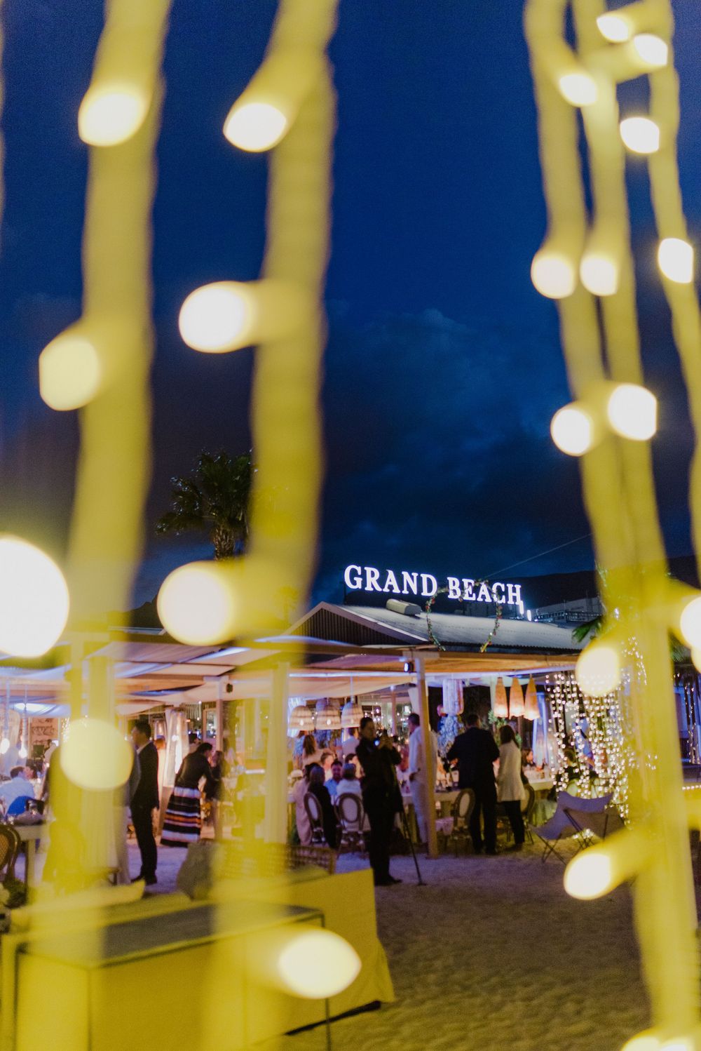 Grand Beach venue at night with glowing string lights bokeh in foreground and guests gathered under illuminated sign.