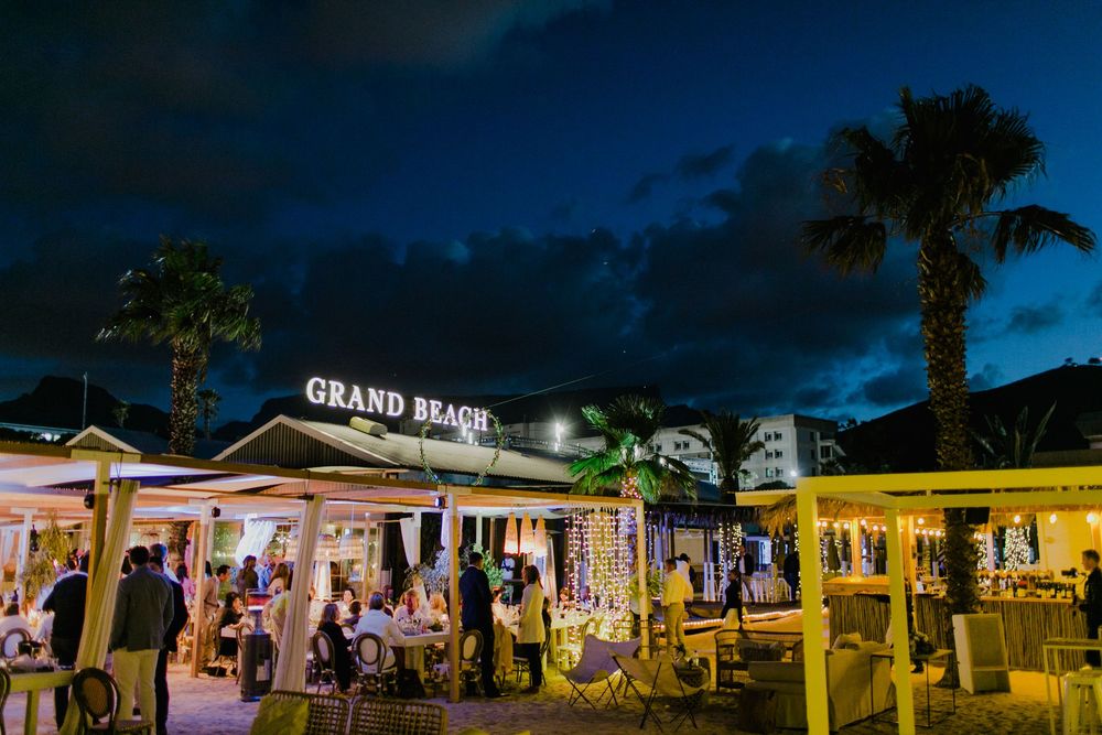 Grand Beach restaurant lit up at night with palm trees, outdoor seating, and a vibrant atmosphere under a dramatic blue sky.