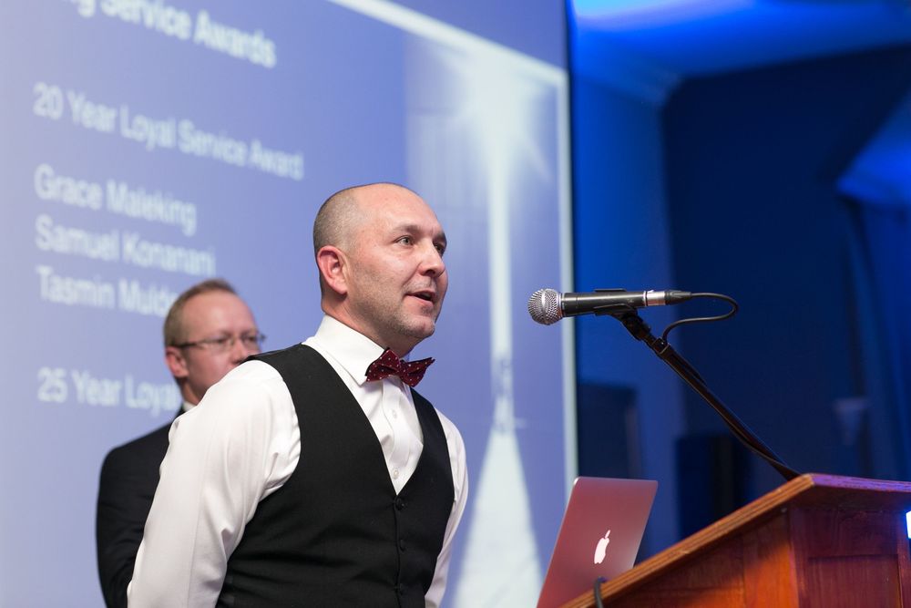Man in vest and bow tie presenting at corporate awards ceremony with projection screen displaying Loyal Service Awards.