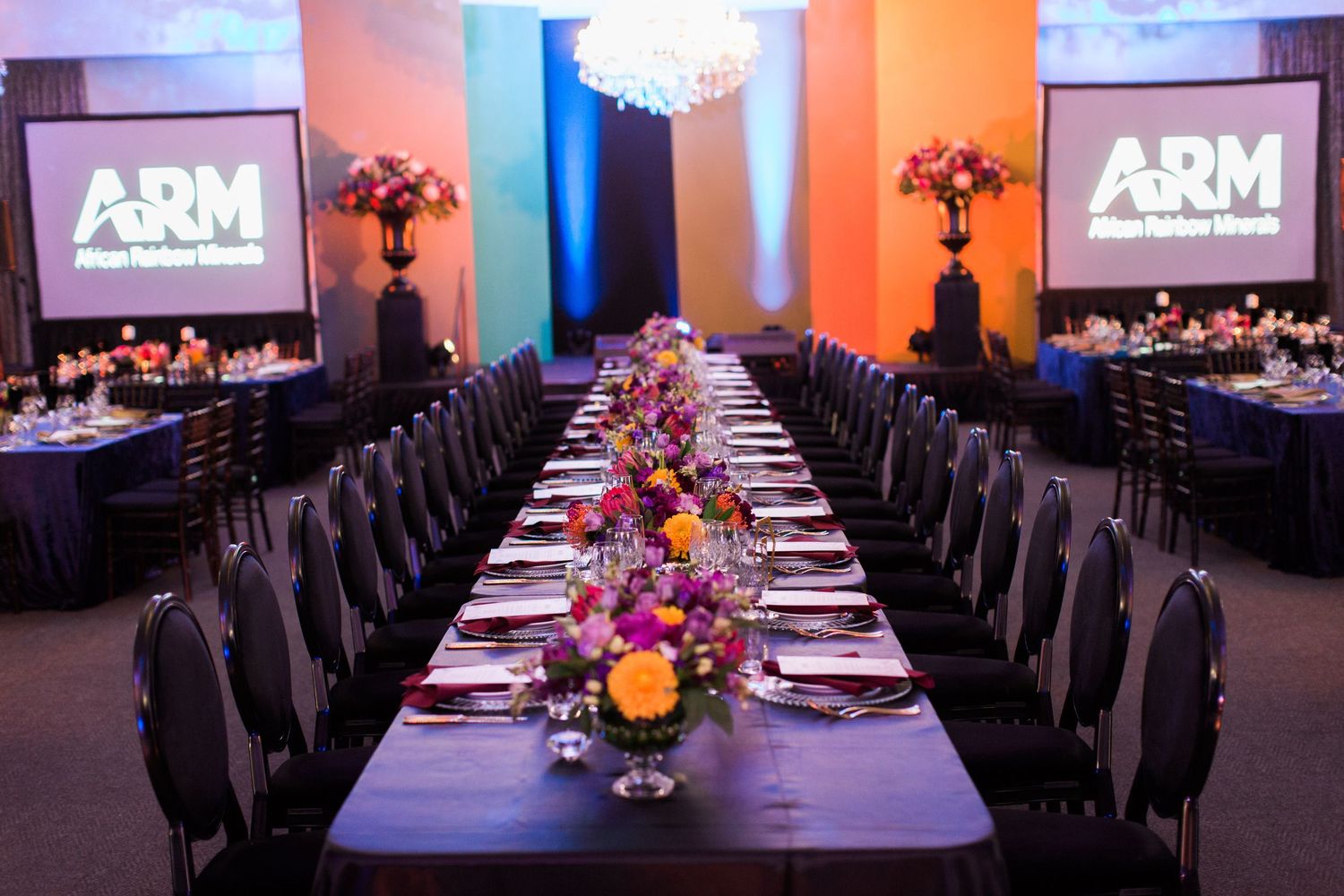 Elegant ARM event banquet hall with long tables, colorful floral centerpieces, navy linens, and branded projection screens.