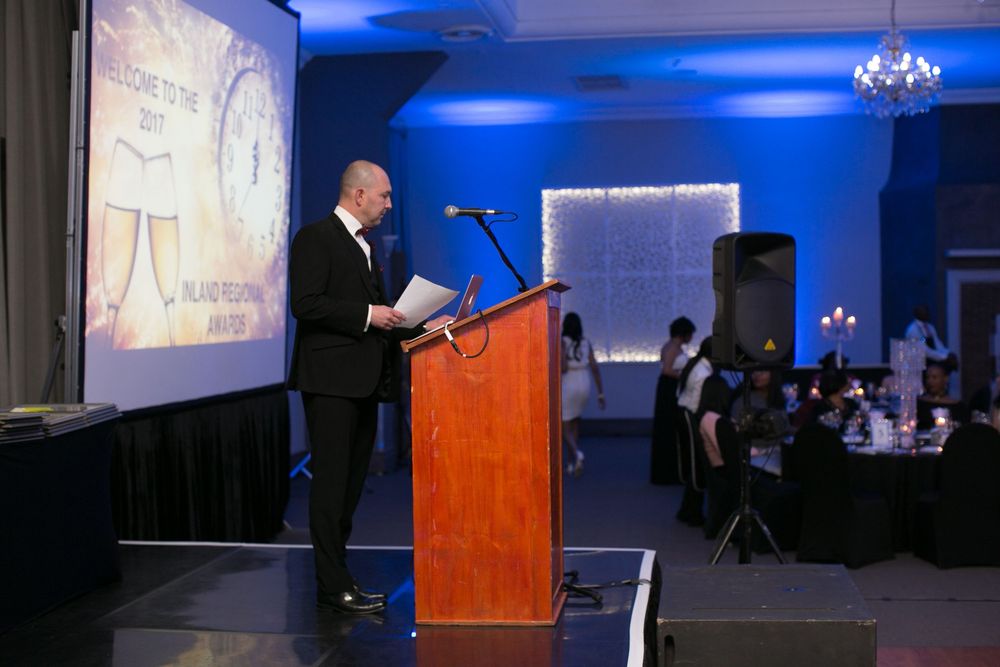 Man speaking at podium during formal awards ceremony with blue ambient lighting and banquet tables in background.