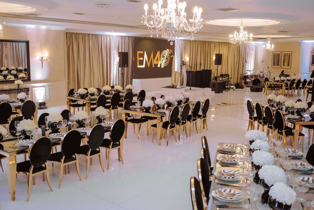 Elegant event hall with black and gold chairs, white floral centerpieces, chandeliers, and EM40 monogram backdrop.