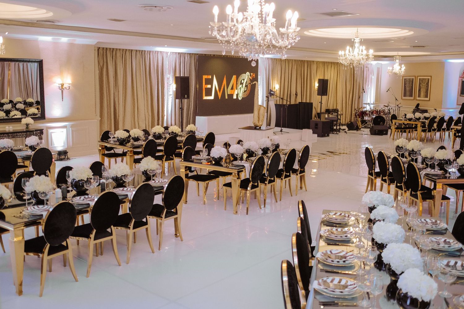 Elegant event hall with black and gold chairs, white floral centerpieces, chandeliers, and EM40 monogram backdrop.