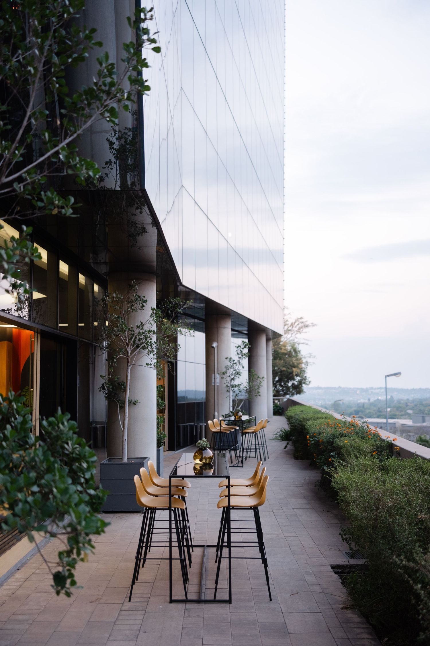 Modern outdoor terrace with yellow bar stools and high tables beside a glass building facade at dusk.