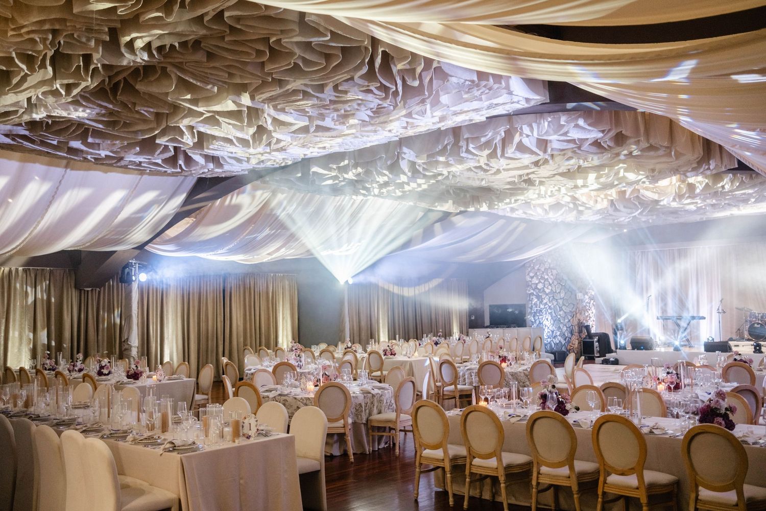 Elegant wedding reception hall with draped white fabric ceiling, fairy lights, round tables, and gold chairs.