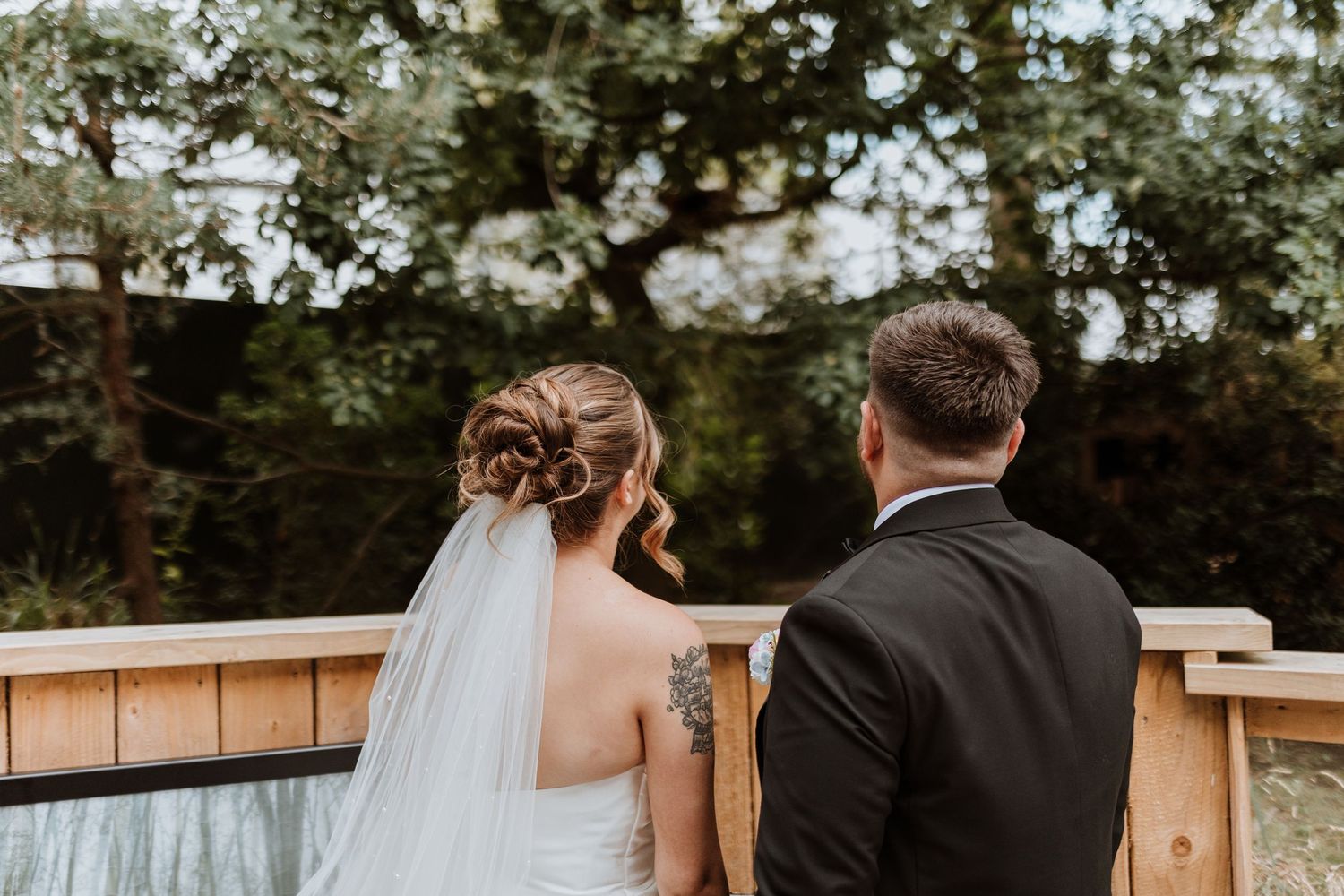 Bride and groom stand together outdoors, bride wearing white gown with veil and floral updo, groom in black suit.
