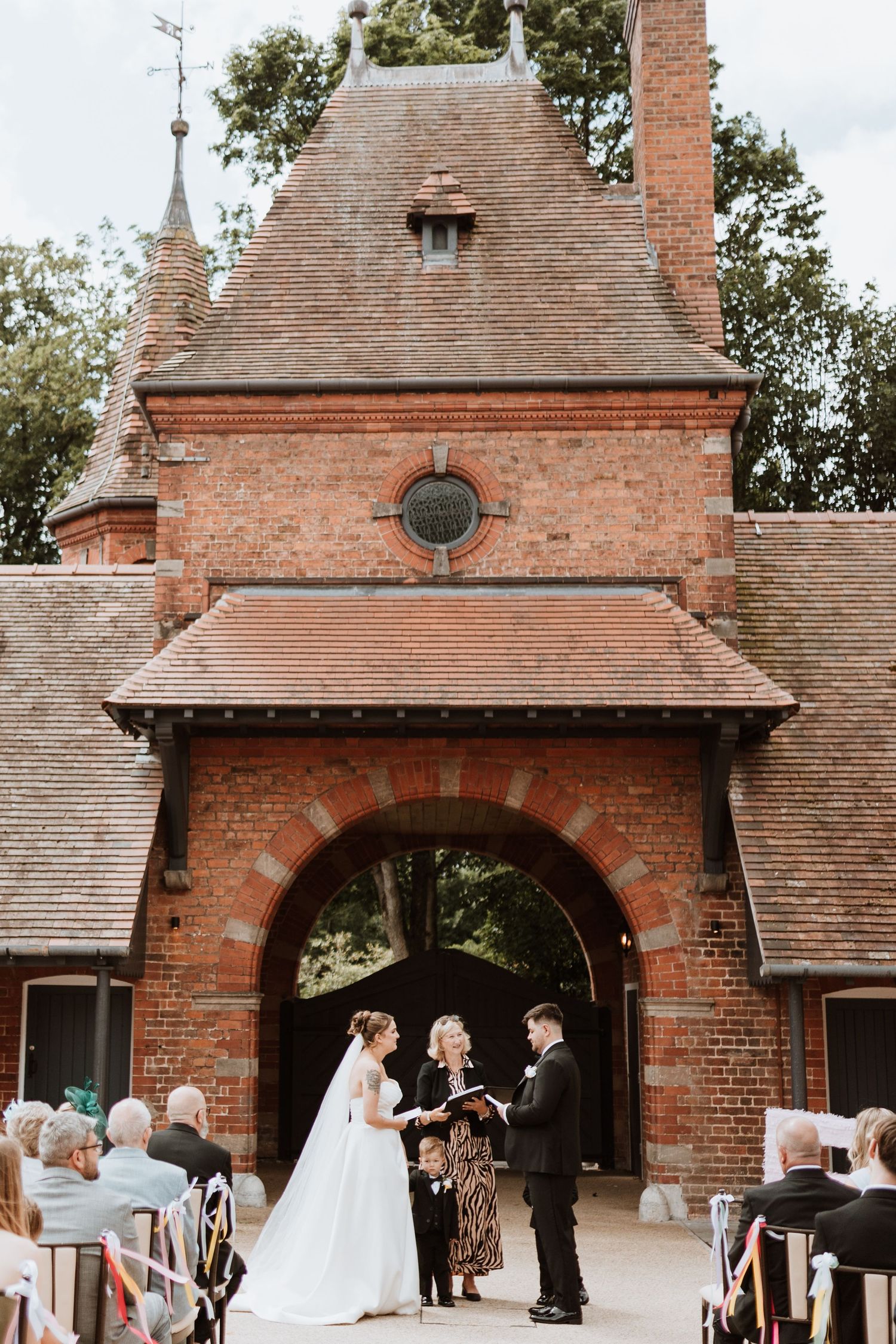 Bride and groom exchange vows before an officiant at an outdoor wedding ceremony in front of a red brick archway.