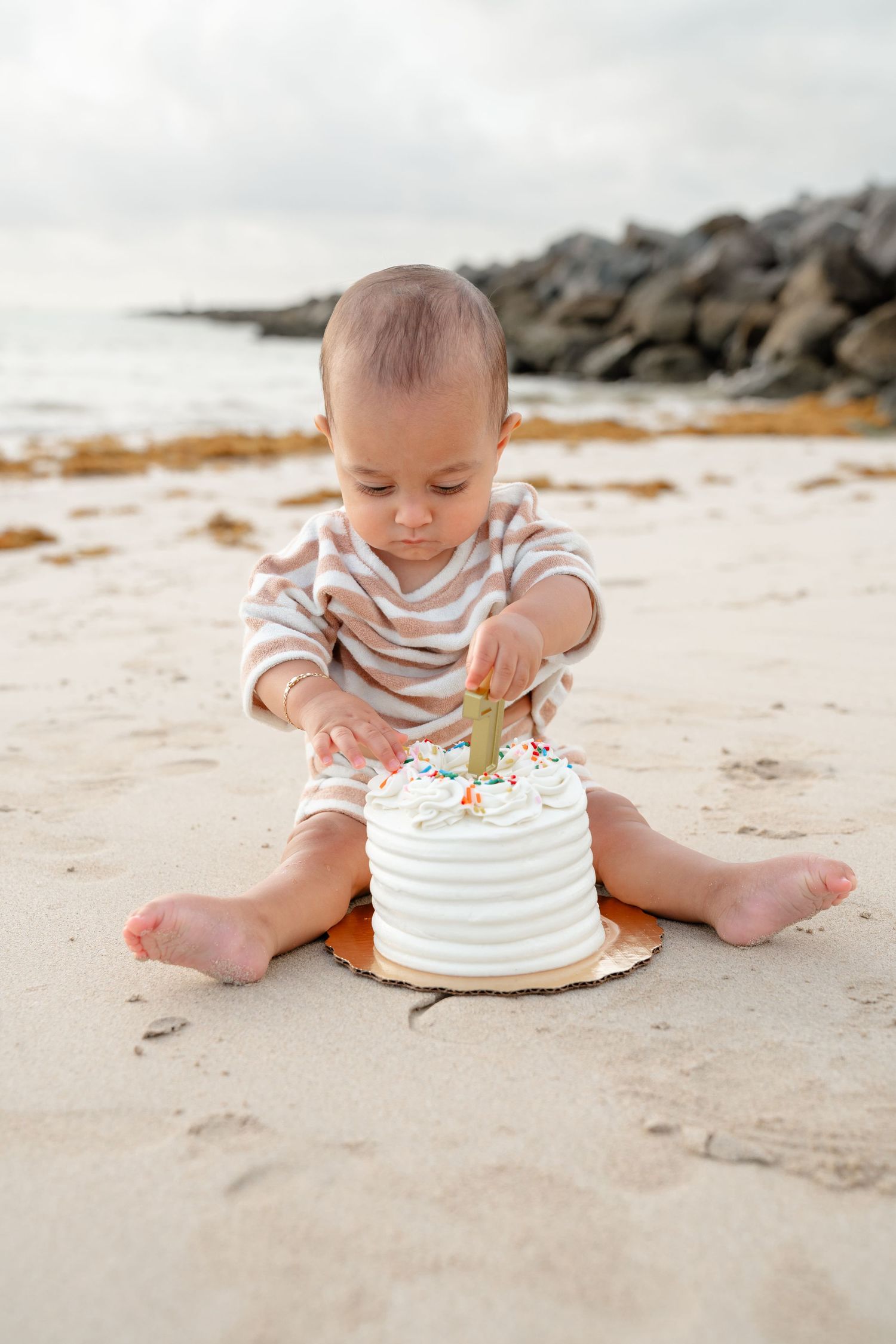 Baby in striped onesie sitting on sandy beach smashing white first birthday cake with colorful sprinkles.