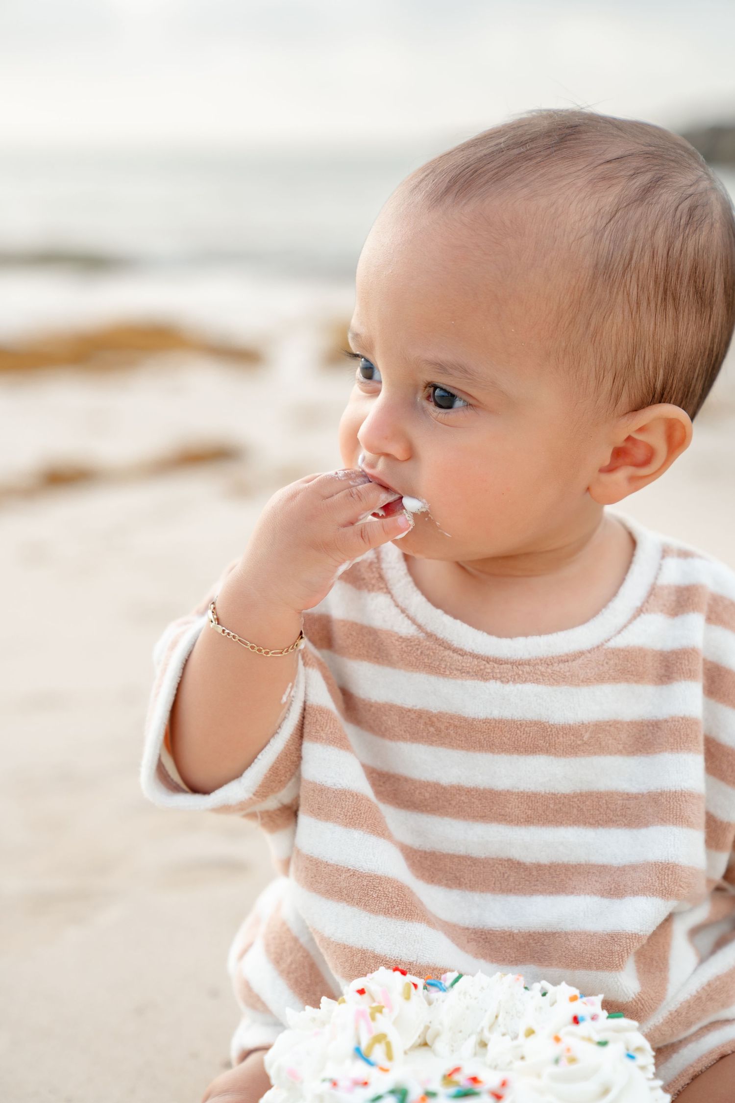 Baby in striped outfit eating cake on beach, celebrating first birthday smash cake session by the ocean.