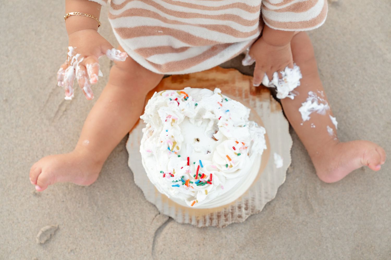Baby sitting on sand with a white frosted birthday smash cake covered in colorful sprinkles during first birthday.