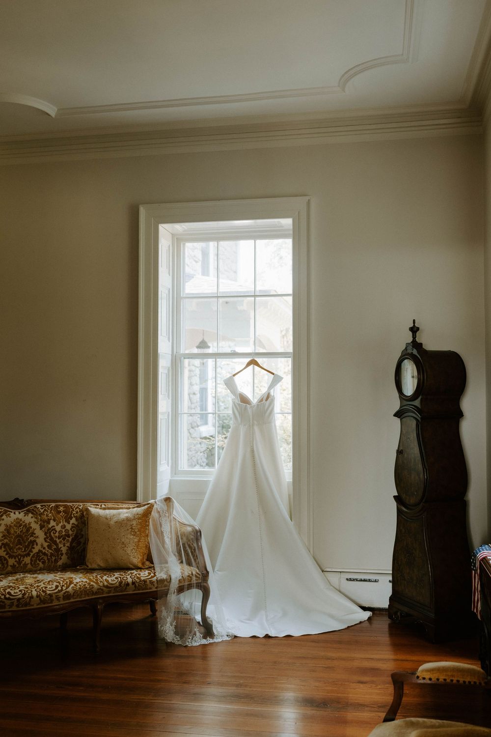 Elegant wedding gown hangs in a window beside a vintage grandfather clock in a classic room.