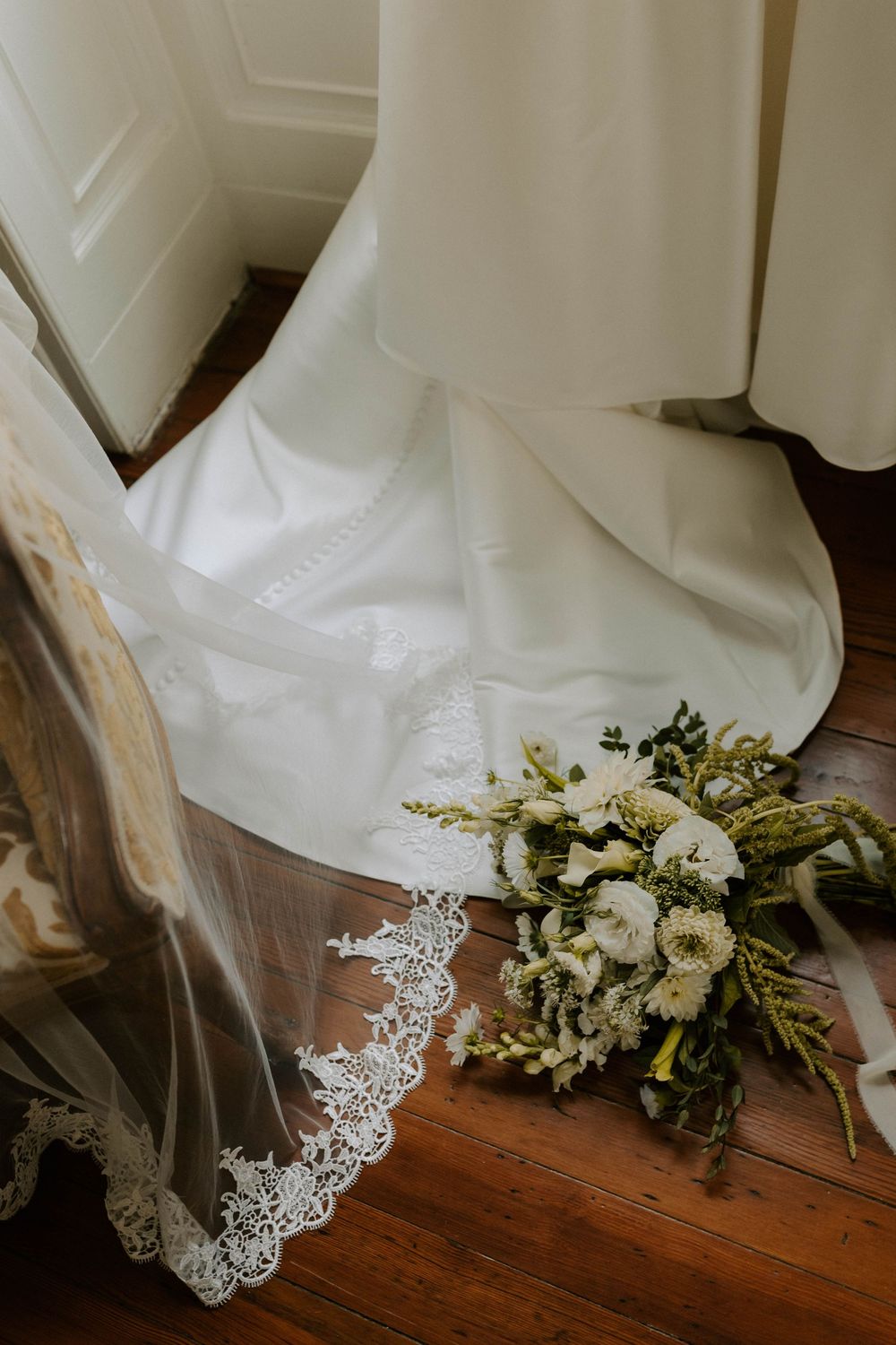 White wedding dress train and lace veil with bridal bouquet on hardwood floor.
