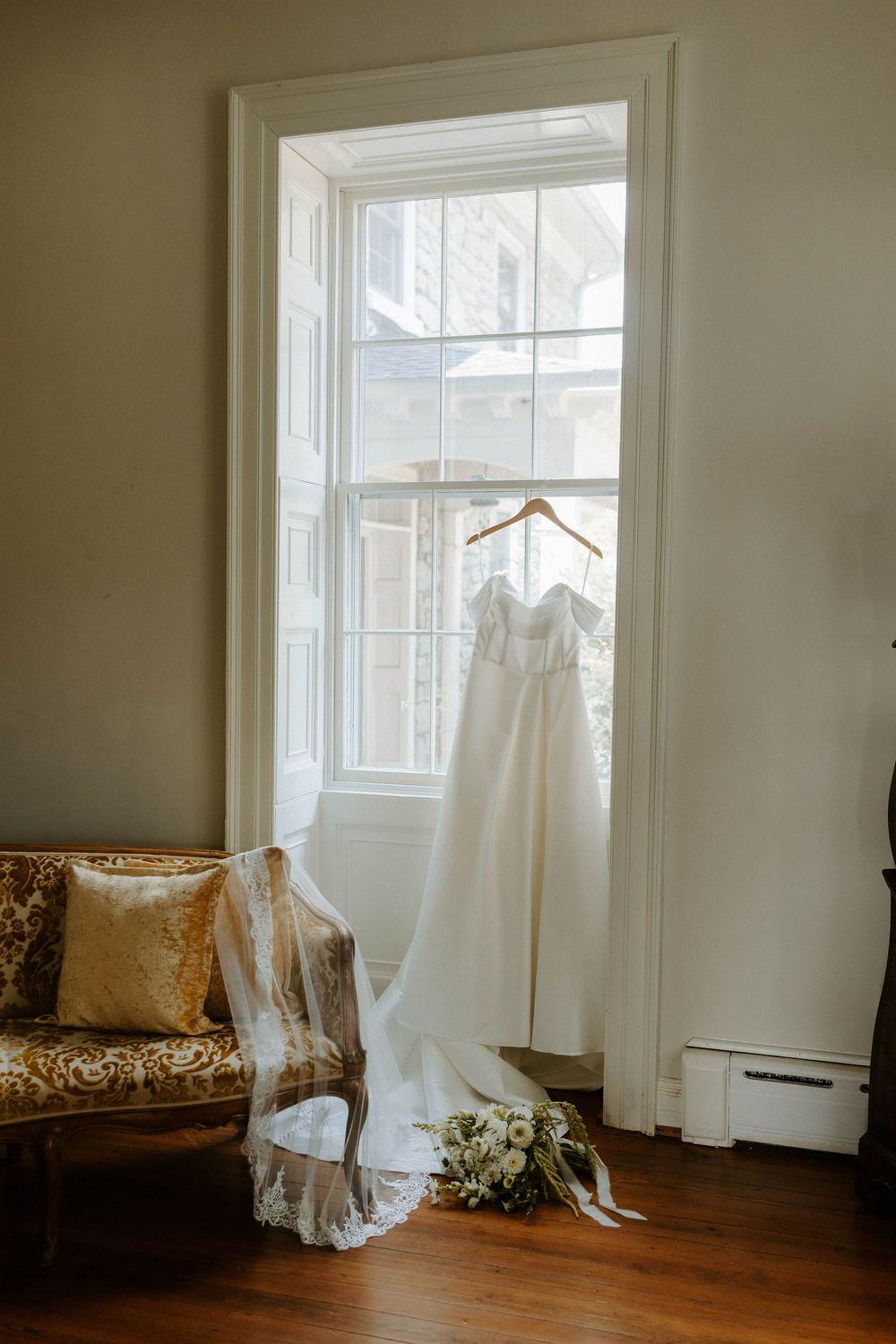 White off-shoulder wedding gown hangs in a sunlit window beside a veil and bridal bouquet on the floor.