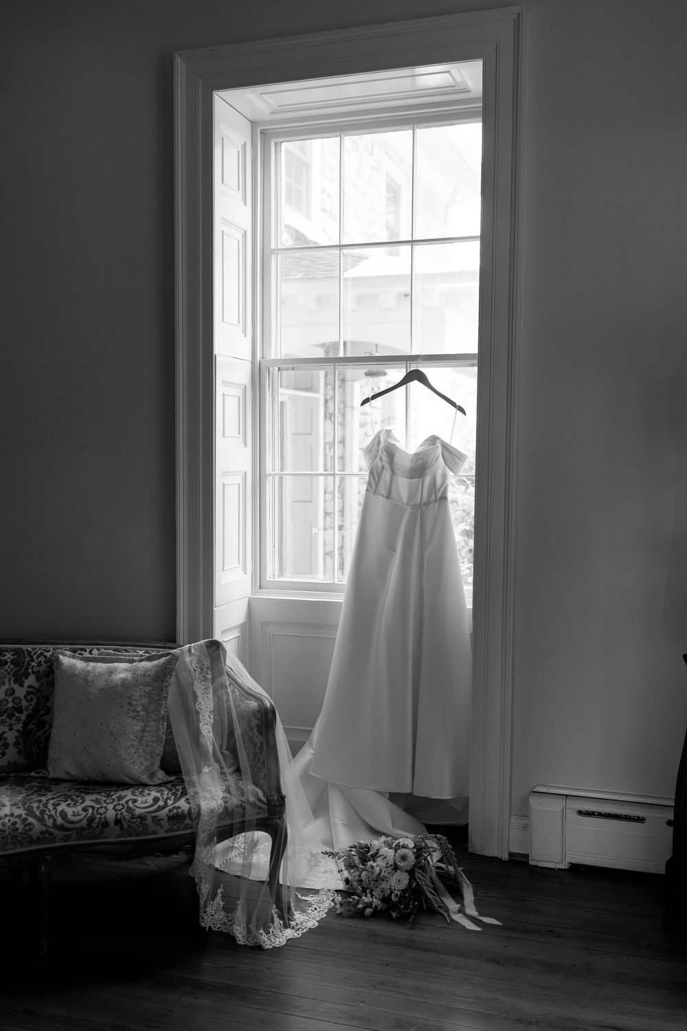 Elegant wedding dress hanging in a window with bridal veil and bouquet in black and white photo.