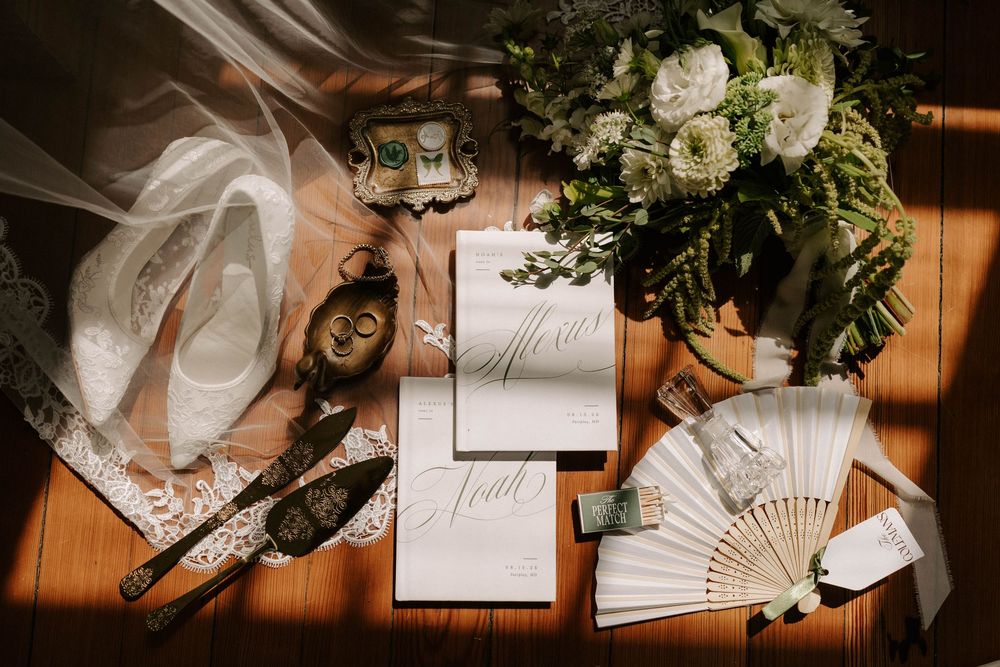 Elegant wedding flat lay featuring white bouquet, invitation suite, fan, shoes, and gold rings on wood surface.