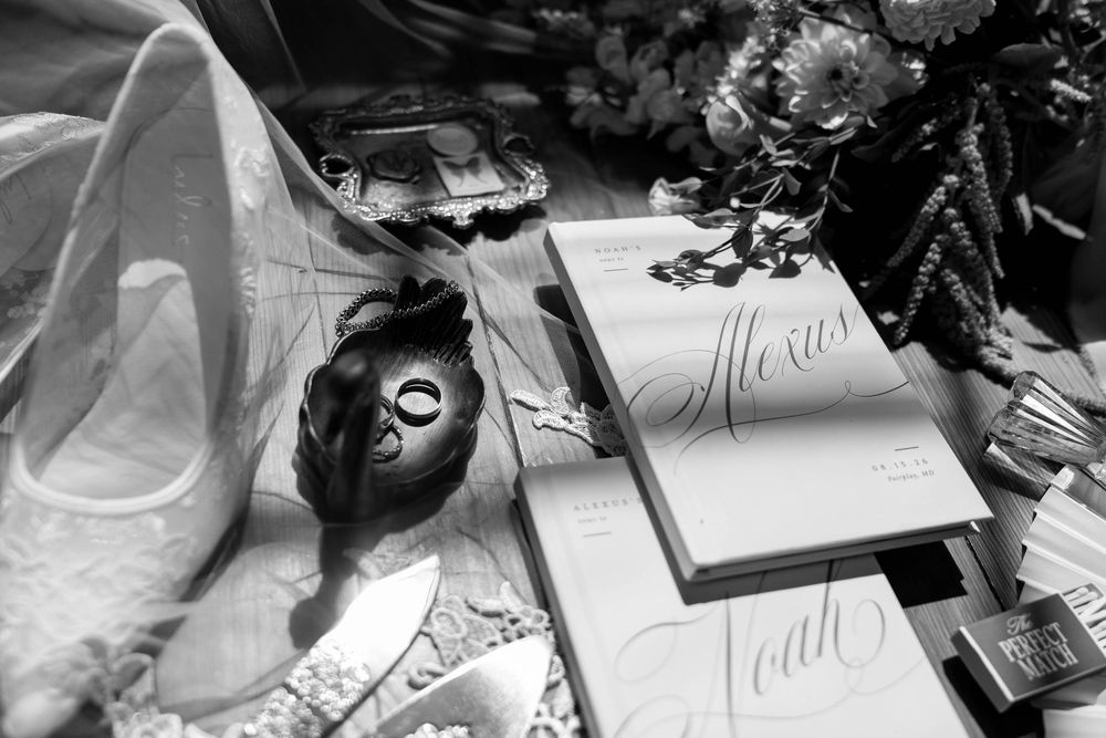 Black and white flat lay of wedding details including shoes, rings, vows, and bridal accessories.