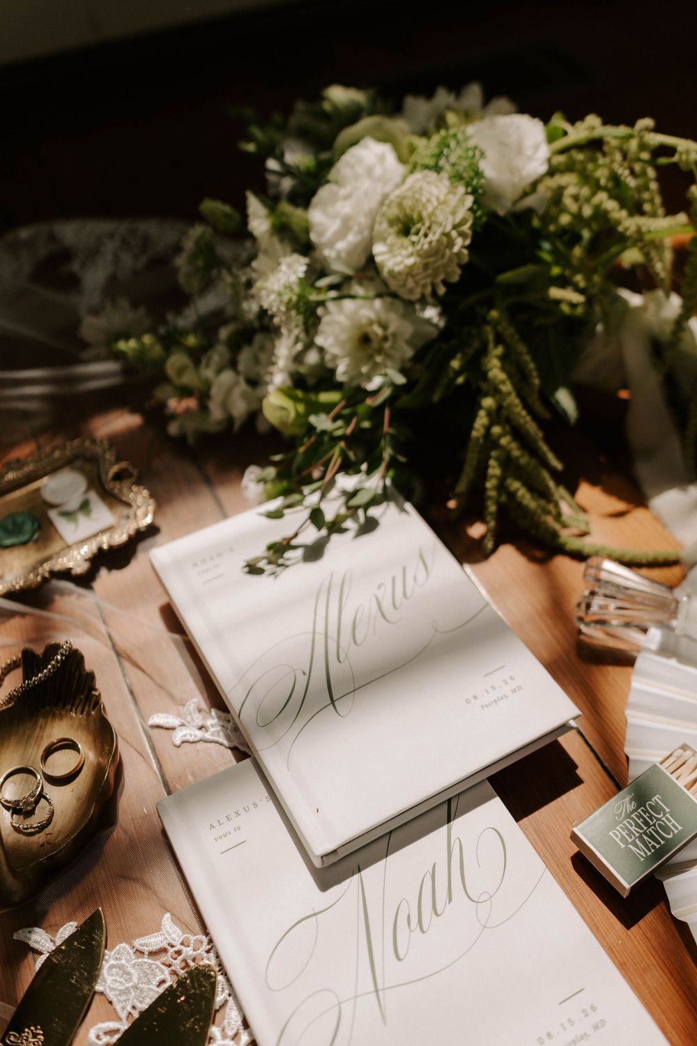 Elegant wedding vow books with calligraphy names beside white floral bouquet and gold rings on wooden table.
