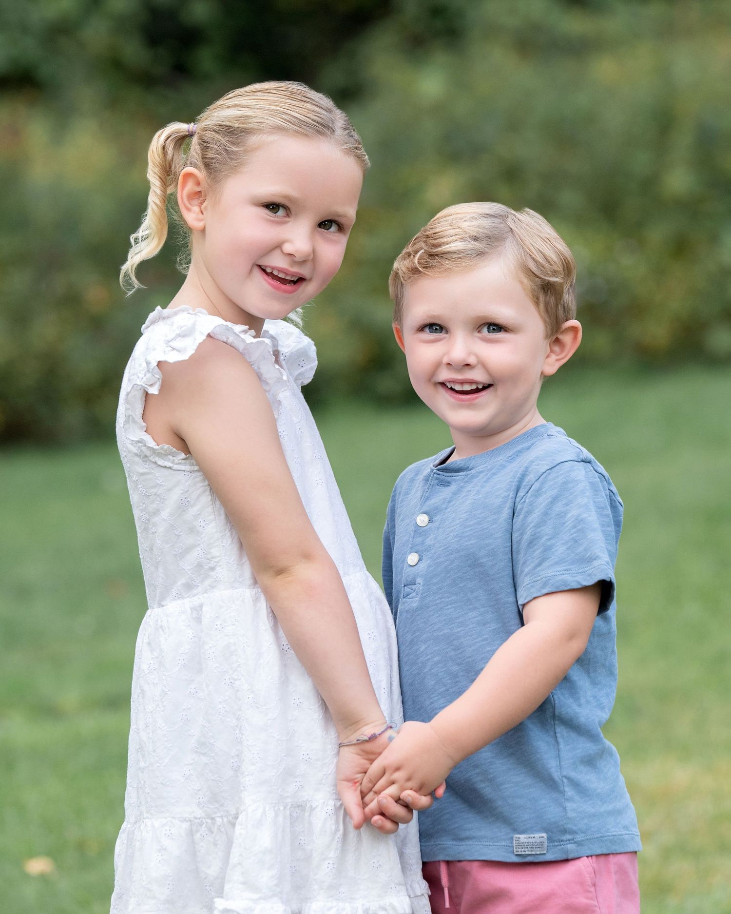 Two smiling children pose back-to-back outdoors, boy in blue suit and girl in blue floral dress with white cardigan.