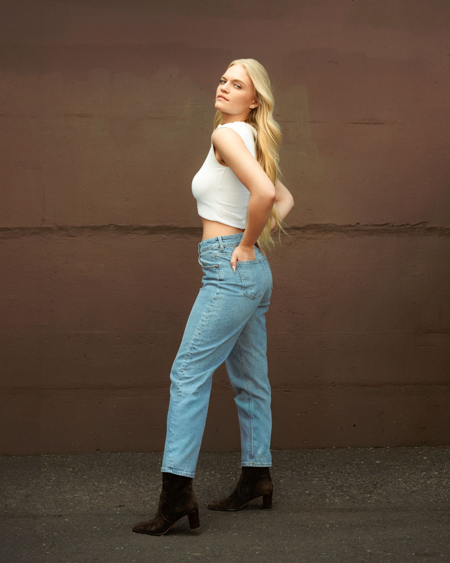 Blonde woman posing in white crop top, light blue jeans, and dark ankle boots against a brown wall.