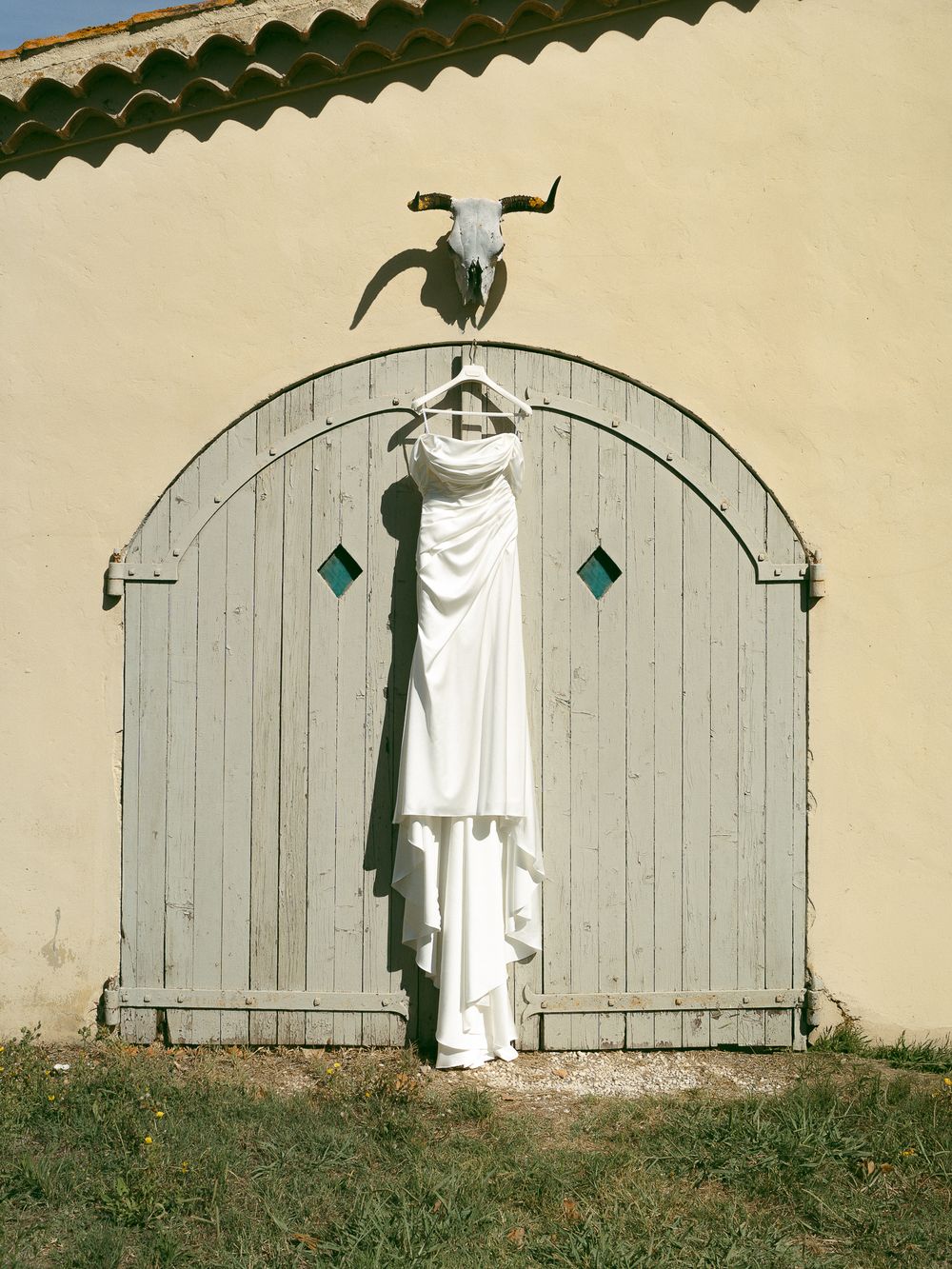 White wedding dress hanging on rustic arched wooden door with bull skull above, against yellow stucco wall.