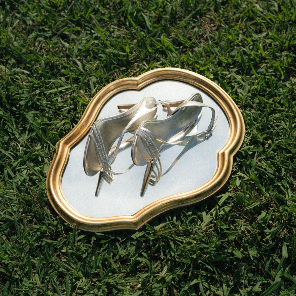 Silver sandal heels displayed on a gold ornate mirror tray placed on lush green grass outdoors.