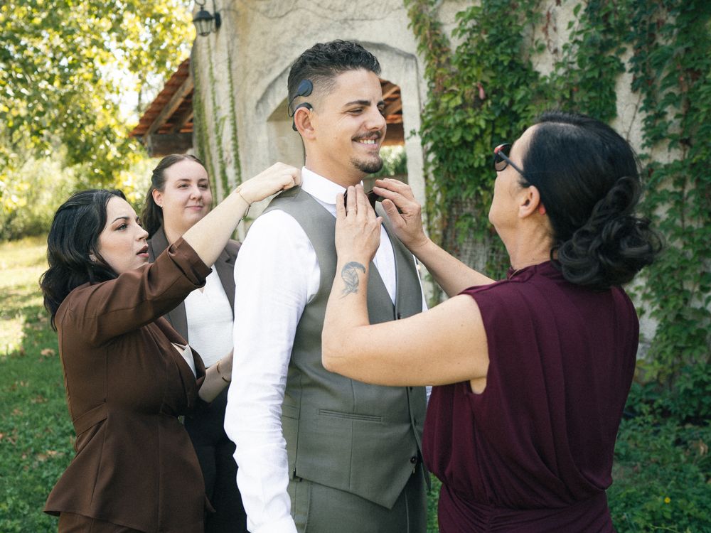 Groom in gray vest being helped by bridesmaids adjusting his tie outdoors at a wedding venue.