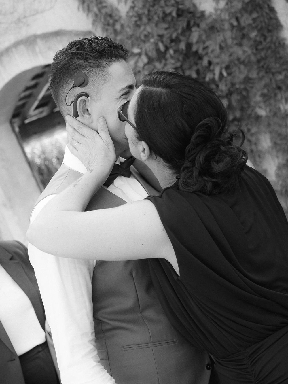 Black and white photo of a woman tenderly kissing a man in a suit at a wedding celebration.