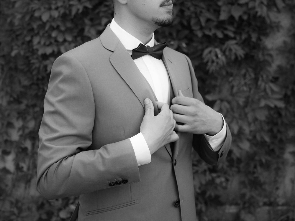 Black and white photo of a well-dressed man in a suit and bow tie adjusting his jacket outdoors.