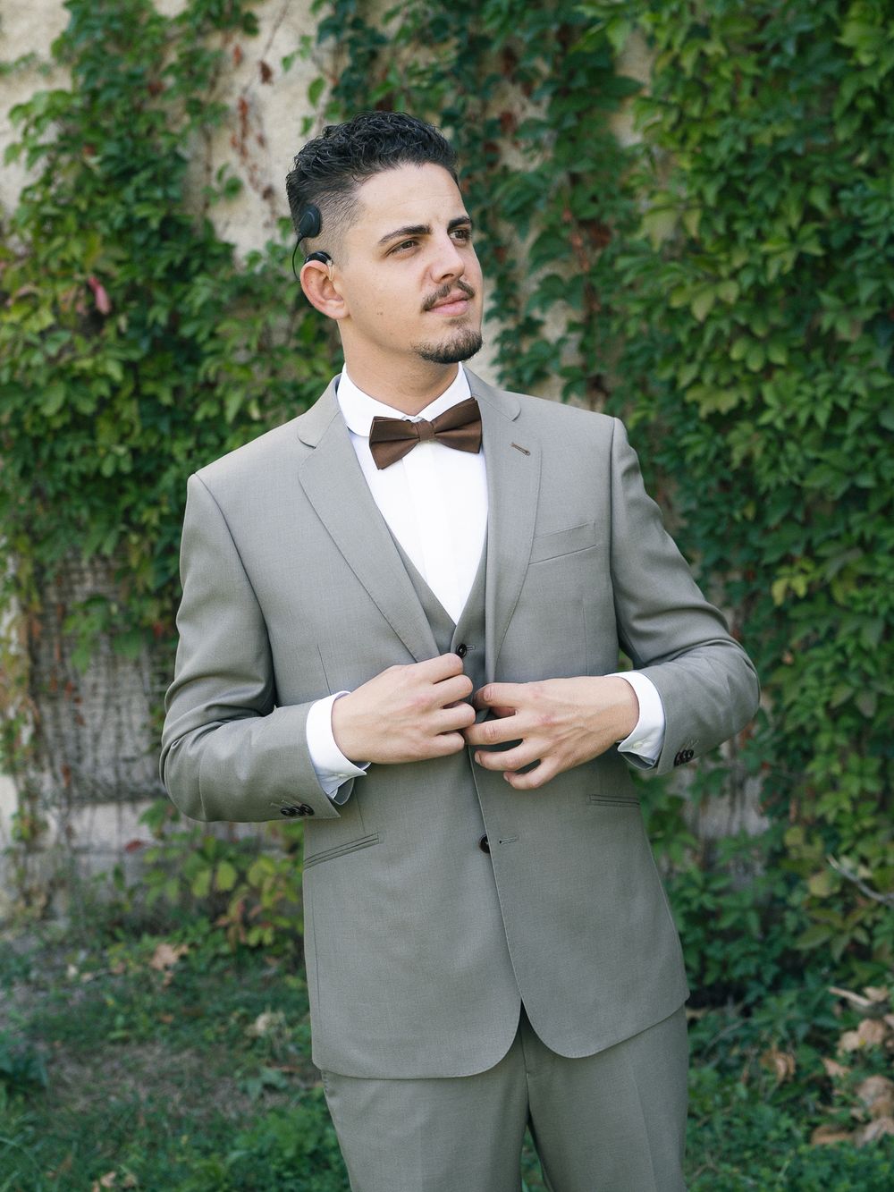 Man in gray three-piece suit with brown bow tie posing outdoors against ivy-covered wall.