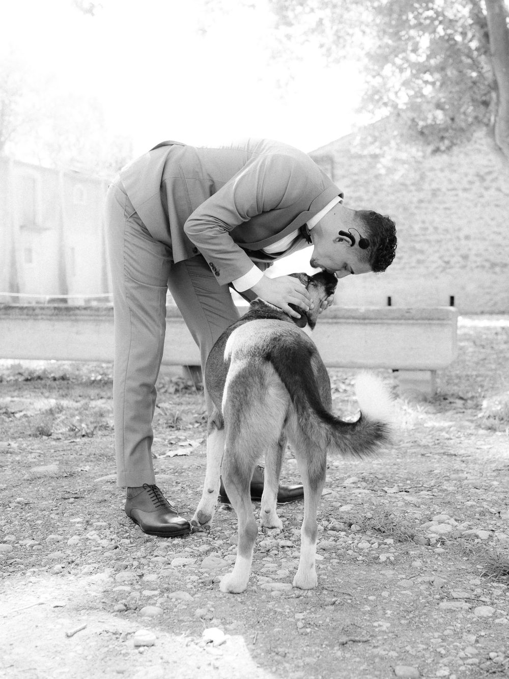 Man in suit bending down to kiss a dog outdoors in a black and white photo.