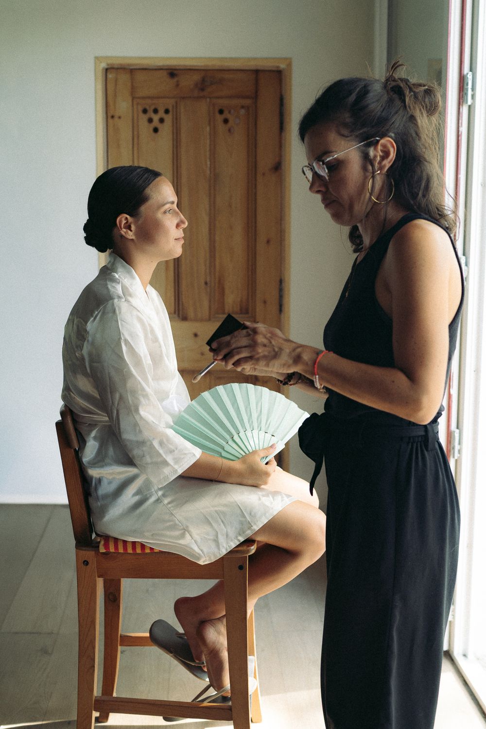 A woman in a white robe sits on a wooden chair holding a mint fan while a stylist in black applies makeup.