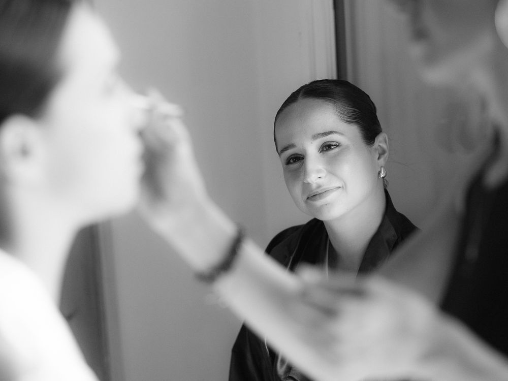 Black and white photo of a woman getting makeup applied by a professional makeup artist in a mirror reflection.