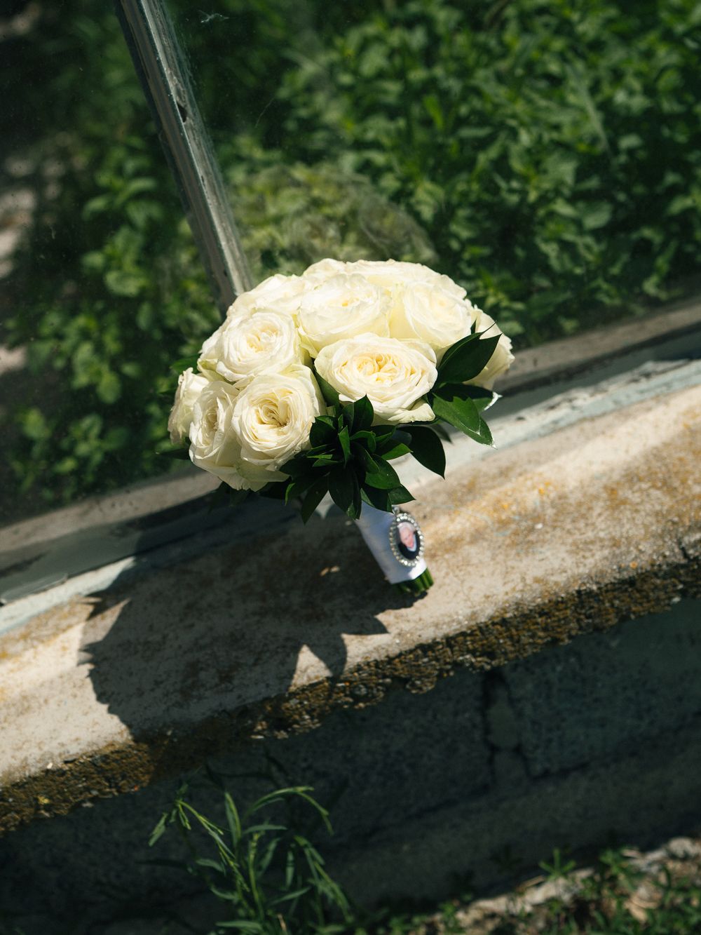 White rose bridal bouquet resting on a window ledge with lush green foliage in the background.