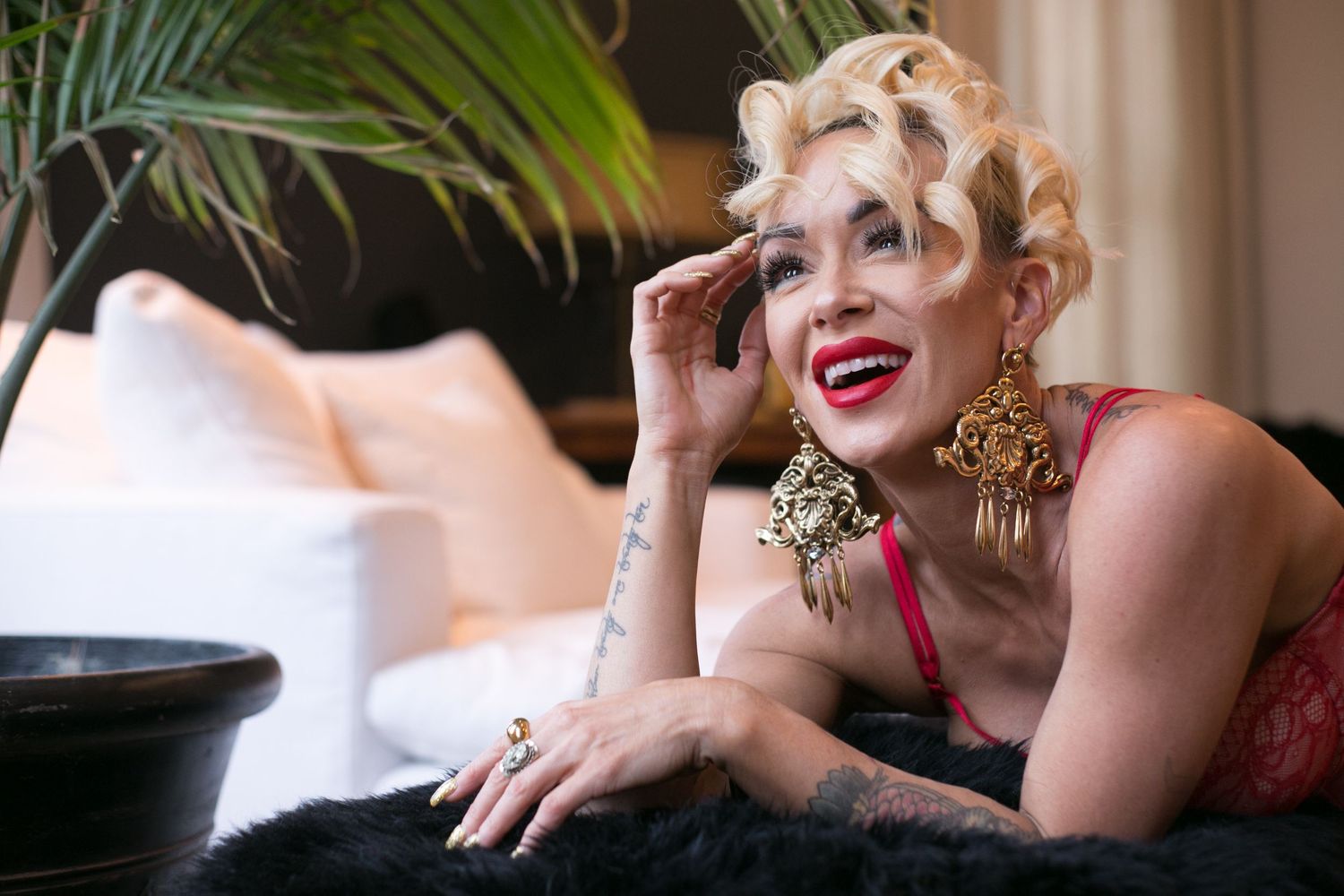 A glamorous blonde woman in red lace lingerie with gold earrings poses elegantly on a black fur surface.