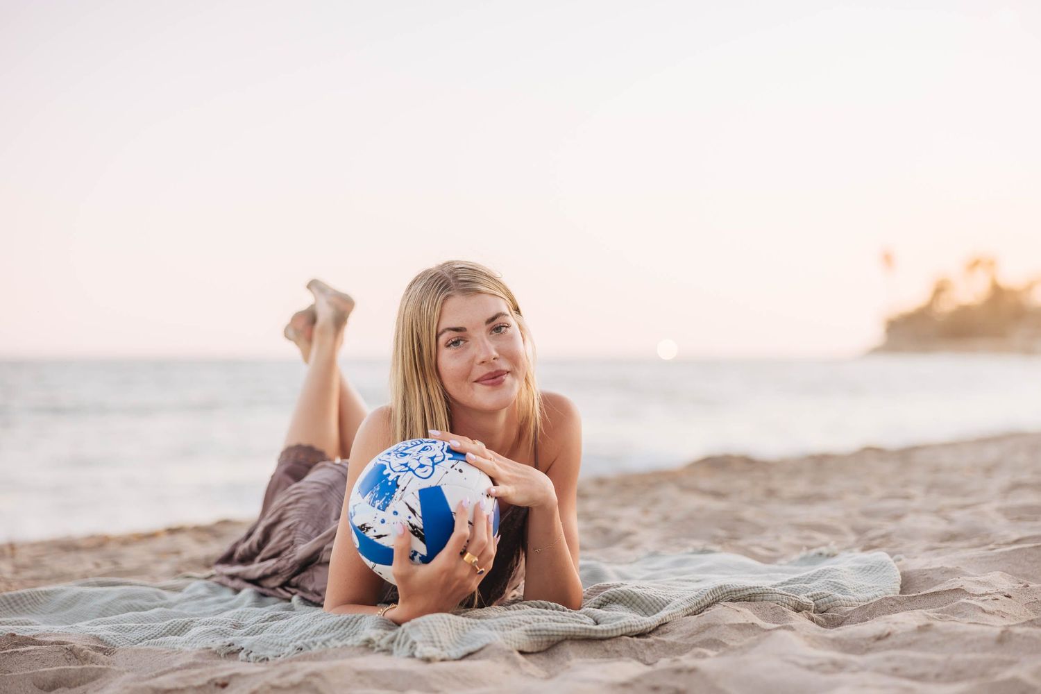 Young blonde woman lying on sandy beach holding a blue and white soccer ball at sunset.