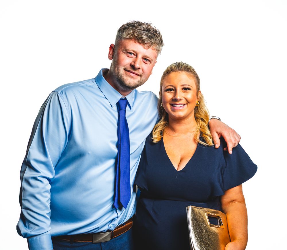 A smiling man in a blue shirt and tie poses with a blonde woman in a navy dress against a white background.