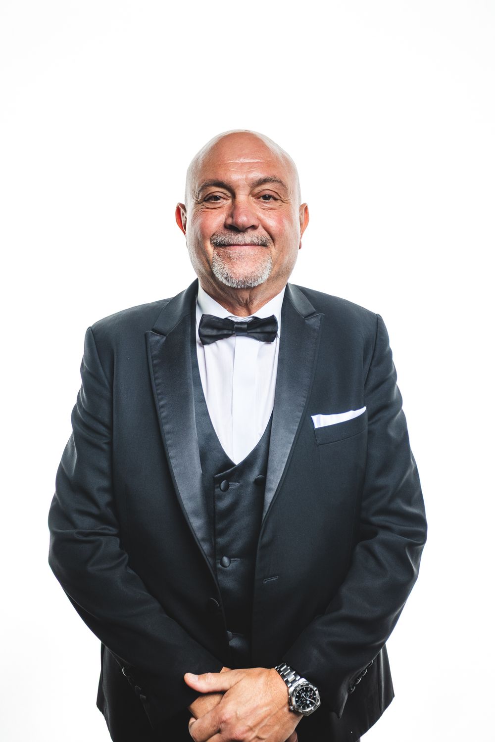 Smiling bald senior man with gray beard wearing black tuxedo with bow tie and watch, posing against white background.