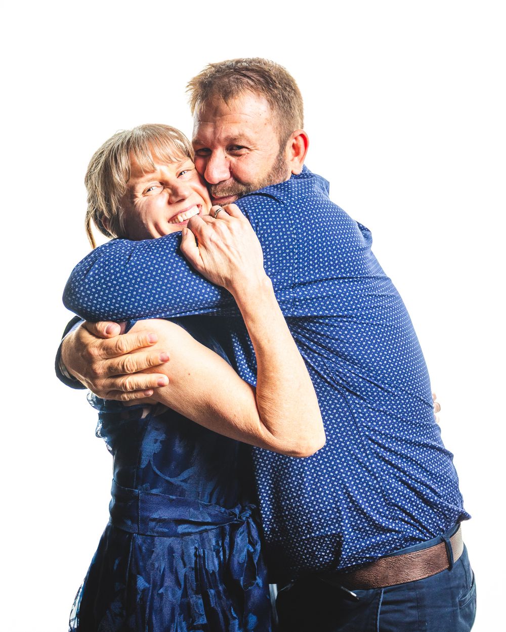An older couple in blue outfits share a warm embrace, smiling joyfully against a white background.
