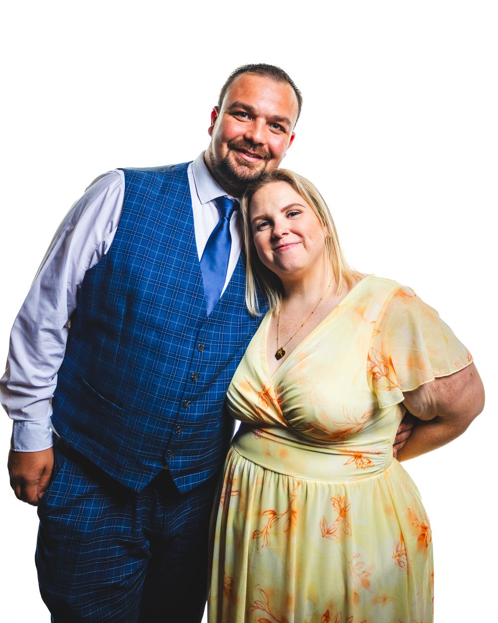 A smiling couple poses together, man in blue vest and tie, woman in yellow floral dress, against white background.