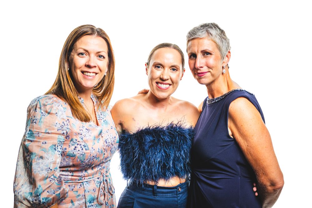 Three smiling women posing together at a formal event, wearing elegant dresses against a white background.
